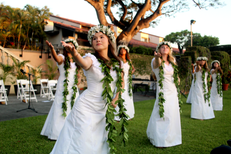 A group of La Pietra students  dressed in white gowns, performing a traditional hula dance on a grassy area.