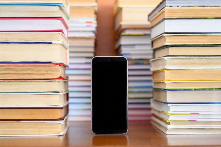 A smartphone with a blank display stands next to tall piles of old books.