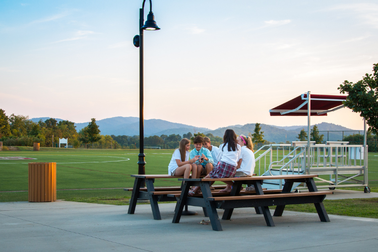 A small group of students chats around a picnic table beside a green sports field.
