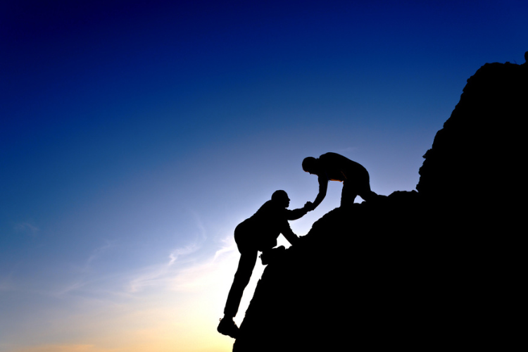 Two silhouetted figures climb a mountain together at sunset