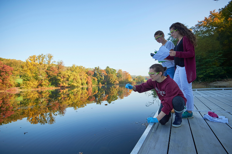 Three students wearing safety goggles collect water samples from a lake's edge.